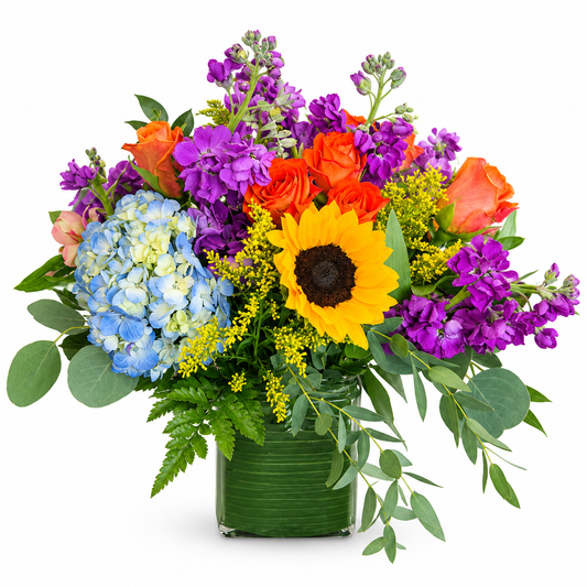 Colorful Bright Moments flower arrangement with sunflowers, blue hydrangeas, orange roses, purple blooms and greenery in a glass vase.